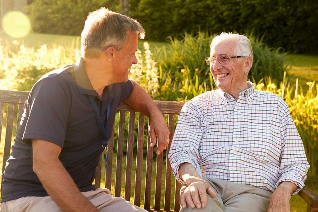 Two older adults sitting together in a bright, comfortable living room, smiling and talking over coffee.