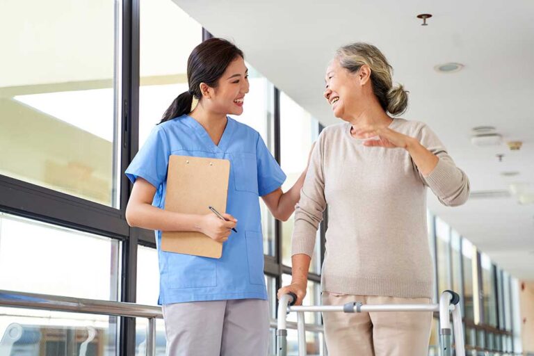 Caregiver sanitizes hands beside an older resident in a bright assisted living common room.